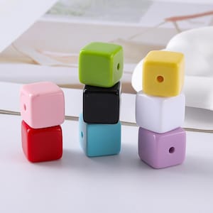 Puede incluir: Una colección de cuentas lisas en forma de cubo en varios colores sólidos, incluyendo rosa claro, rojo, verde lima, negro, azul brillante, amarillo, blanco y morado claro. Cada cuenta presenta un único orificio central para enhebrar, con acabado brillante.