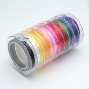 Puede incluir: Un recipiente cilíndrico de plástico transparente que contiene una pila de múltiples carretes de cordón fino en un arcoíris de colores que incluye negro, blanco, amarillo, naranja, rosa, rojo, verde, azul y morado, dispuestos en columna.