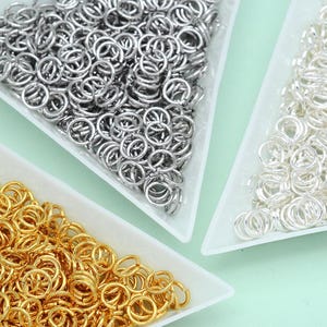 May include: A collection of metal jump rings organized in three white triangular trays. The assortment includes dark silver-toned, bright silver-toned, and gold-toned circular rings, each featuring a small split for jewelry assembly.