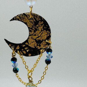 Puede incluir: Collar de cadena dorada con un colgante de media luna negro y dorado, un cristal azul claro y cuentas de cristal azules y negras.