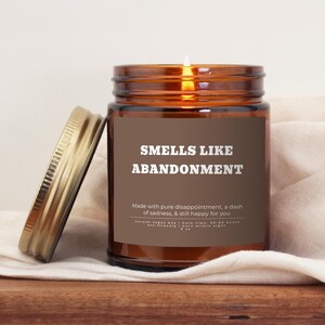 May include: A brown glass jar candle with a gold lid, labeled "SMELLS LIKE ABANDONMENT." The label also reads "Made with pure disappointment, a dash of sadness, & still happy for you." The candle sits on a wooden surface.