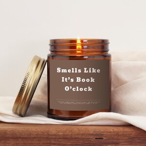 May include: A brown glass candle jar with a gold lid. The candle is lit and the label reads "Smells Like It's Book O'clock".