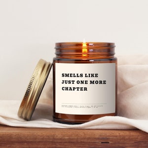 Puede incluir: Un tarro de vela de vidrio marrón con una tapa dorada. La vela está encendida y el tarro tiene una etiqueta blanca con el texto "Smells like just one more chapter".
