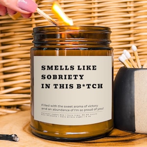 May include: A brown glass candle with a white label that reads "Smells Like Sobriety In This B*tch". The label also includes the text "Filled with the sweet aroma of victory and an abundance of I'm so proud of you!"