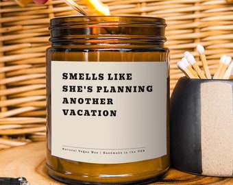 Puede incluir: Una vela de vidrio marrón con una etiqueta blanca que dice "Smells like she's planning another vacation". La vela está encendida y hay una cerilla sobre una superficie de madera junto a ella.