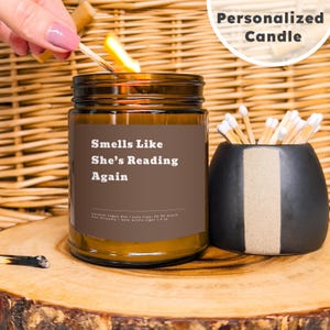 Op de afbeelding: Een bruine glazen kaars met een bruin label waarop "Smells Like She's Reading Again" staat. De kaars is aangestoken en er is een lucifer op de achtergrond.