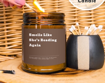 May include: A brown glass candle with a brown label that reads "Smells Like She's Reading Again". The candle is lit and there is a matchstick in the background.