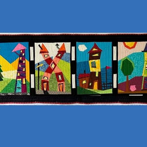Puede incluir: Un tapiz rectangular de tela con cuatro paneles distintos de texturas acolchadas. Cada panel representa una escena de casa colorida con formas geométricas, incluyendo una torre morada, una estructura en forma de X y una casa marrón.