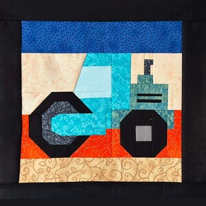 May include: A colorful quilt block featuring a tractor design. The tractor is primarily light blue and teal, with black wheels and accents. The background includes orange, beige, and blue patterned fabrics, all set against a black border.
