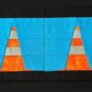 May include: A fabric art piece featuring two orange and gray traffic cones on a bright blue background. The cones are set against a blue backdrop with horizontal lines, framed by a black border. The design is a patchwork style.