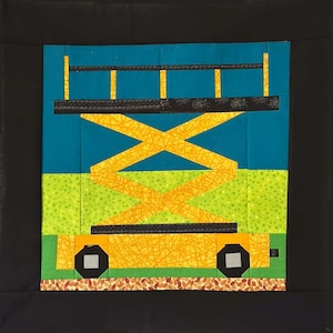 May include: A colorful quilt block depicting a yellow scissor lift against a blue sky and green field. The lift has black wheels and a black top. The background is a black square.
