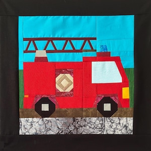 May include: A quilted artwork depicting a red fire truck with a blue sky background. The fire truck has a ladder, a light blue window, and black wheels. The artwork is framed in black.