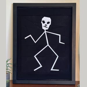 May include: Framed artwork with a white geometric stick figure against a black backdrop. The figure has a skull-like head. The artwork is in a black frame, creating a stark contrast. The artwork is a modern, minimalist design.