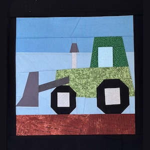 May include: A patchwork quilt square featuring a green and black bulldozer with a gray blade and smokestack. The background is blue, and the ground is brown. The quilt is framed in black.