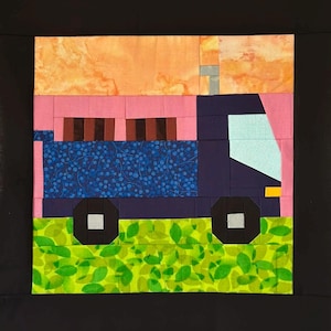 May include: A colorful quilt block featuring a stylized truck. The truck is primarily navy blue with a blue patterned bed, pink cab, and orange top. The wheels are black, and the ground is green with leaf patterns. The background is black.