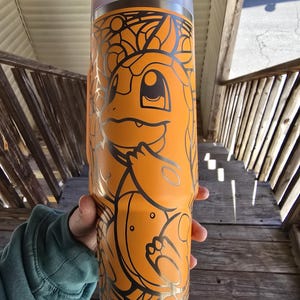 Puede incluir: Vaso naranja con un diseño de Charmander negro. El vaso de acero inoxidable tiene una tapa blanca y una pajita. El diseño de Charmander es una ilustración estilizada. El vaso es cilíndrico.