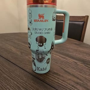 Może przedstawiać: Jasnoniebieski kubek termiczny ze stali nierdzewnej z czarnym uchwytem. Kubek przedstawia różne postacie ze studia Ghibli, w tym Totoro, Bez Twarzy i Koci Autobus. Kubek ma również logo Stanley i napis "Studio Ghibli".