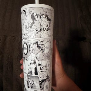 Könnte beinhalten: Ein weißer zylindrischer Becher mit Strohhalm, der mit einem schwarz-weißen Comic-Druck im Manga-Stil versehen ist. Das Design umfasst mehrere Panels mit Charakteren, Sprechblasen mit englischem Text wie GRAB, GRRT und HA sowie Actionszenen.