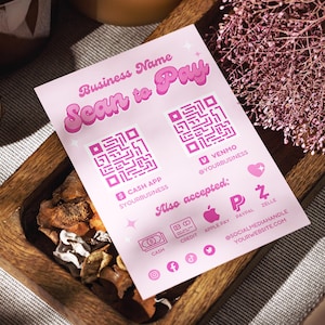 May include: A pink business card with the text "Scan to Pay" and QR codes for Cash App and Venmo. The card also lists accepted payment methods like Apple Pay, PayPal, and Zelle. The card is in a wooden tray.