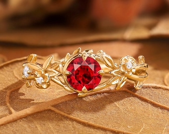 May include: A gold ring featuring a large, round, red gemstone at the center, surrounded by a floral design. Small, clear accent stones are set in the floral elements. The ring is set on a brown leaf.