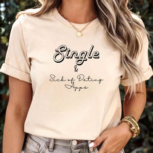 以下が含まれることがあります： 黒い筆記体で「Single & Sick of Dating Apps」と書かれたベージュ色の半袖Tシャツ。「Single」の文字は太字で縁取りされています。柔らかい素材のシャツです。