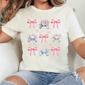 May include: A white t-shirt with a graphic design of pink bows and floral game controllers.