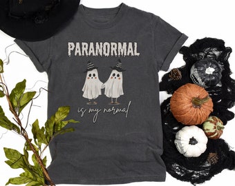 May include: A charcoal gray t-shirt featuring a graphic of two white ghosts wearing black witch hats and striped socks, holding hands. The text above reads PARANORMAL in a dripping font, and the text below reads is my normal in script.