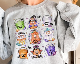 May include: A light gray sweatshirt featuring a colorful Halloween-themed graphic. The graphic includes various cartoon characters such as pumpkins, ghosts, and bats, all in a cute and playful style. The design incorporates candy corn and other festive elements.