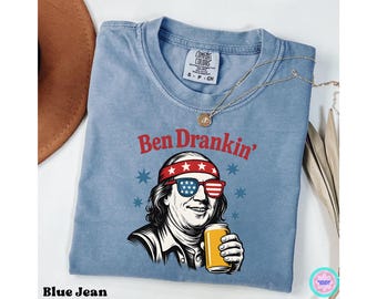 Puede incluir: Camiseta azul vaquero con un gráfico de Benjamin Franklin con gafas de sol y una diadema con estrellas. El texto "Ben Drankin'" está encima del gráfico. Un collar dorado está drapeado sobre la camiseta.