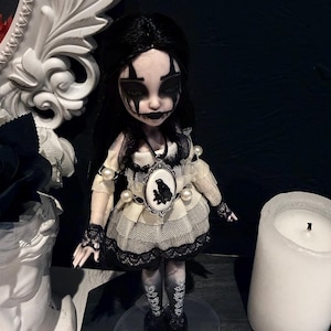 Puede incluir: Muñeca gótica con cabello negro y maquillaje dramático. La muñeca viste un vestido color crema con detalles de encaje negro y un colgante de camafeo. Se encuentra sobre un soporte transparente, con una gran vela blanca al fondo.