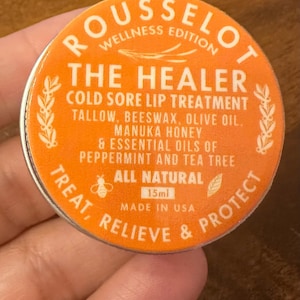 May include: Orange round tin with a white label that reads "ROUSSELOT WELLNESS EDITION THE HEALER COLD SORE LIP TREATMENT TALLOW, BEESWAX, OLIVE OIL. MANUKA HONEY & ESSENTIAL OILS OF PEPPERMINT AND TEA TREE ALL NATURAL 15ml MADE IN USA TREAT, RELIEVE & PROTECT"
