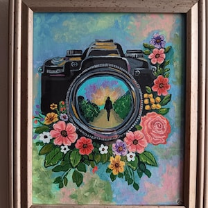 May include: A framed painting featuring a black camera surrounded by colorful flowers. The camera lens shows a person walking down a path. The background has a mix of blue, pink, and green hues.