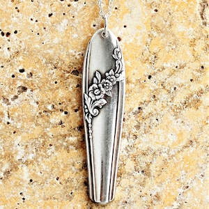 May include: A silver pendant necklace with a floral design. The pendant is an elongated oval shape, with a detailed floral pattern on one side. The necklace chain is also silver and is attached to the pendant at the top.