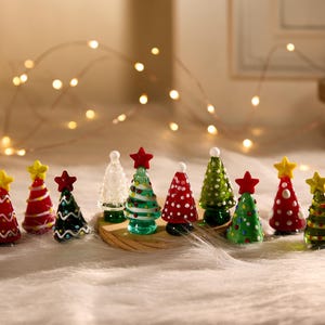 May include: A selection of miniature Christmas tree ornaments in red, green, and white, each with a star on top. The trees are displayed on a soft, white surface, with fairy lights in the background, creating a festive scene.