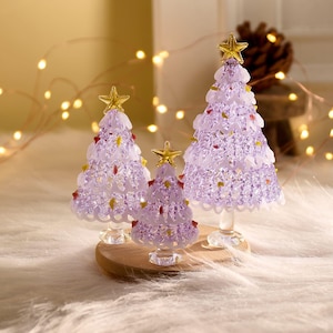 以下が含まれることがあります： さまざまなサイズの装飾的なクリスマスツリーが3つ。ツリーはラベンダー色で、透明なベースと金色の星のトッパーが付いています。ツリーは小さな赤と黄色のアクセントで飾られています。木製のベースにあります。
