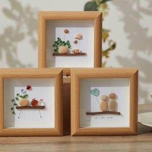 Puede incluir: Tres obras de arte enmarcadas con guijarros. Cada una presenta un diseño único de guijarros y vidrio, que representa escenas como flores y figuras. Los marcos son cuadrados, de madera clara, y están sobre una superficie de madera. Una pieza incluye la fecha 13/8/2015.