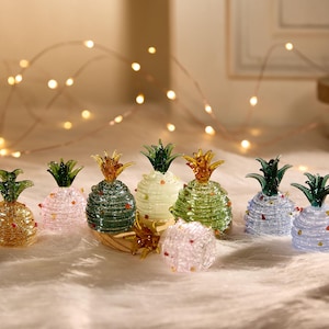 以下が含まれることがあります： 金、ピンク、緑、青など、さまざまな色のガラス製パイナップルの置物のコレクション。各パイナップルは、緑色のトップと小さなカラフルなアクセントを備えた詳細なデザインです。置物は、柔らかい白い表面に配置されています。