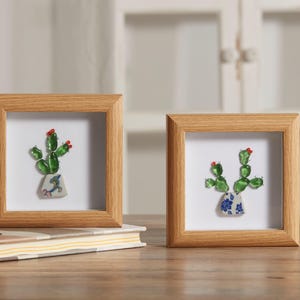 May include: Two framed artworks featuring glass cactus sculptures. The cacti are green with red accents, potted in decorative ceramic pots. The frames are square, made of light-colored wood, and set against a white background.