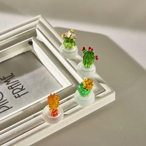 Op de afbeelding: Een witte fotolijst met de tekst "PHOTO FRAME" is versierd met vier miniatuur glazen cactussculpturen. Elke cactus heeft levendige kleuren, waaronder groen, geel en rood, en staat in een kleine, heldere pot. De lijst staat op een lichtgekleurd oppervlak.