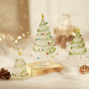 May include: Three glass Christmas trees of varying sizes, decorated with green, white and red beads. The largest tree is on a wooden base and the smallest tree is on a white surface.
