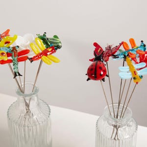May include: Two glass vases containing decorative garden stakes shaped like insects. The stakes depict ladybirds, dragonflies, and butterflies in red, yellow, and blue. The vases are clear with a ribbed design.