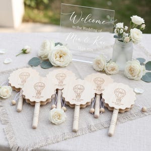 May include: A clear acrylic wedding sign with white text reading Welcome to the Wedding of Mia & Kai 12.18.2026, accompanied by six round wooden hand fans featuring a hot air balloon graphic, the text Just Married, and Up, Up, and Away.