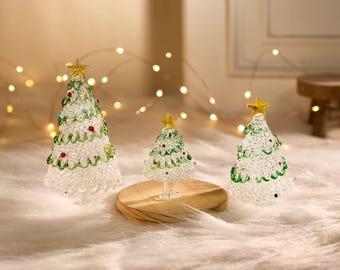 May include: Three decorative glass Christmas trees. Each tree is clear with green and red accents, topped with a gold star. The trees are of varying sizes, with the smallest one on a wooden base.