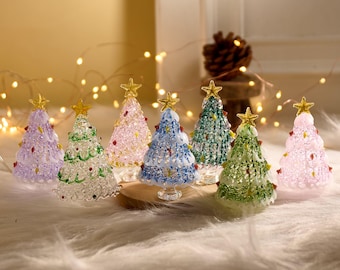 May include: A collection of miniature Christmas trees in various colors, including purple, green, pink, and blue. Each tree is adorned with decorative elements and topped with a gold star. The trees are arranged on a wooden base.