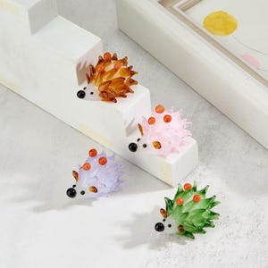 May include: Four glass figurines of hedgehogs in different colors: brown, pink, purple, and green. The hedgehogs are arranged on a white tiered display stand.