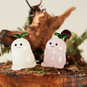 以下が含まれることがあります： 緑色の葉っぱと笑顔の顔をした、ラディッシュの形をした小さなガラスの置物2つ。左側の置物は白で、右側の置物はピンク。緑の苔が生えた木の表面の上に置かれている。