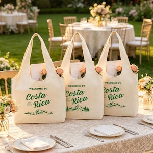 Puede incluir: Tres bolsas de tela color hueso con texto verde que dice Welcome to Costa Rica y Est 2026, junto con un gráfico verde de montañas y bosque. Cada bolsa contiene una tarjeta blanca y una rosa color melocotón dentro de la abertura principal.