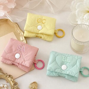 May include: Three quilted earbud cases in pastel yellow, pink, and mint green. Each case features a flap with a white snap button, embroidered initials, and a matching circular plastic ring. The yellow case has GC, the pink has SA, and the mint has BL.