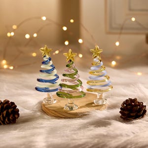 May include: Three decorative glass Christmas trees with gold star toppers. The trees are blue, green, and yellow, with a spiral design. They sit on a wooden base with two pine cones nearby. Fairy lights are in the background.