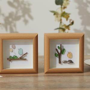 May include: Two framed art pieces with wooden frames. Each frame contains a small, colorful bird made of pebbles. One bird is perched on a branch with balloons, the other near a tree. The artwork is displayed on a wooden surface.